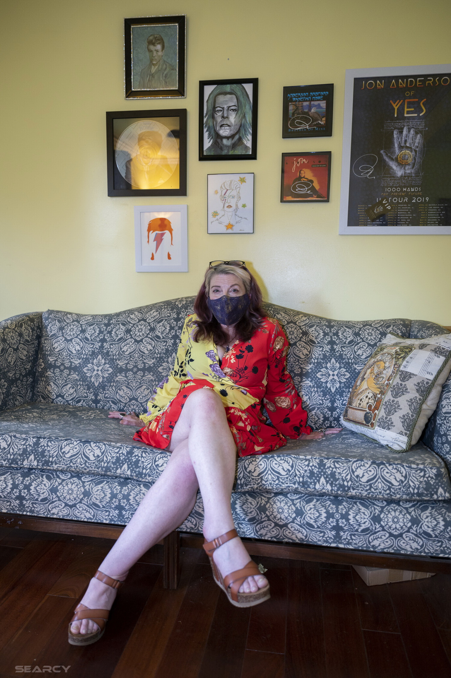 Alicia Search sitting on a blue and white couch wearing a blue mask and yellow and red flowery dress. She has her legs crossed and wearing sandals. on the wall behind her lots of David Bowies posters and prints.
