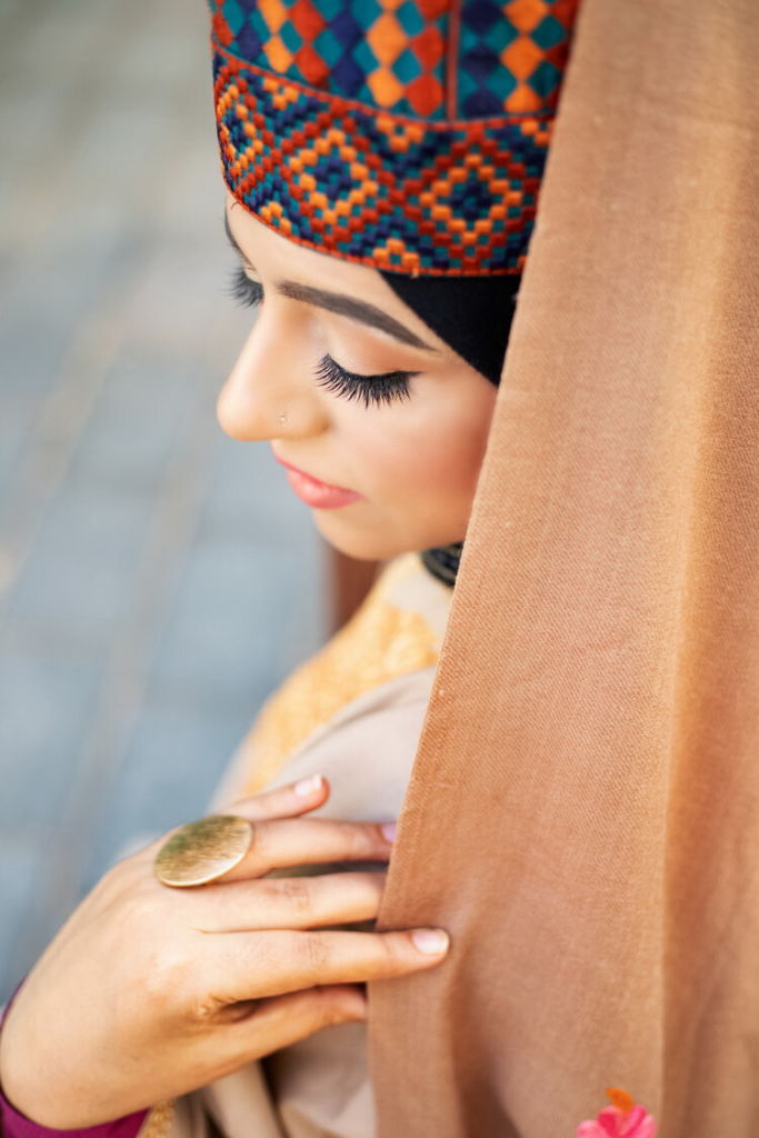 Asiya, lady wearing a headgear in different colors in a geometrical pattern. The shot is from above and her head is covered by a hijab (veil) in rose gold. She is holding the veil with 2 fingers and wearing a gold roud ring.