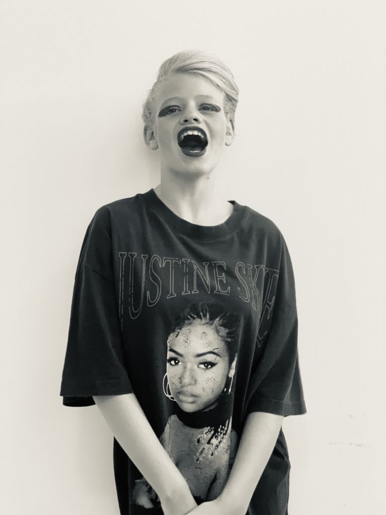 Clara Woods a white teenager with blonde hair tied up, wearing black eyeliner and lipstick. She has her mouth open like in a laugh. She is wearing a black t-shirt with a black woman on it. Her arms are crossed in the front.