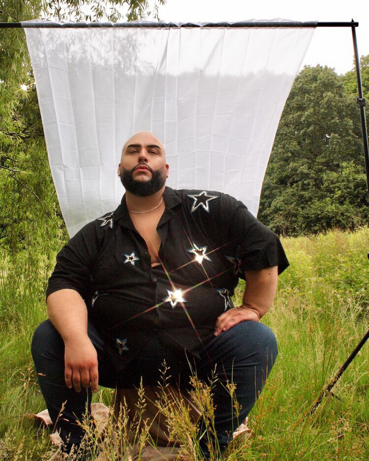 Rural atmosphere, green grass and trees. Ady, the big, plus, latinx model is sitting on a stool with his left hand on one knee and right arm rested on the other. His is looking confidently at the camera, has bold head and black short and full beard He is wearing a black shirt with silver stars and dark denim. Behind him stands a white drape.