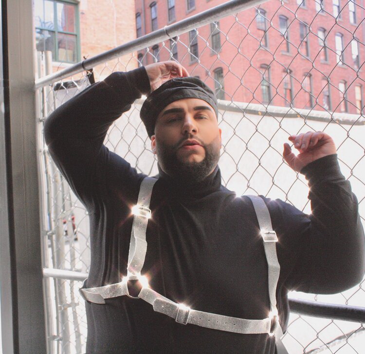 Ady is standing against a net wall, with his both arms lifted, right more stretched up than the other. He is wearing a black turtleneck jumper and a silver suspenders connected by a silver belt across his bust.
