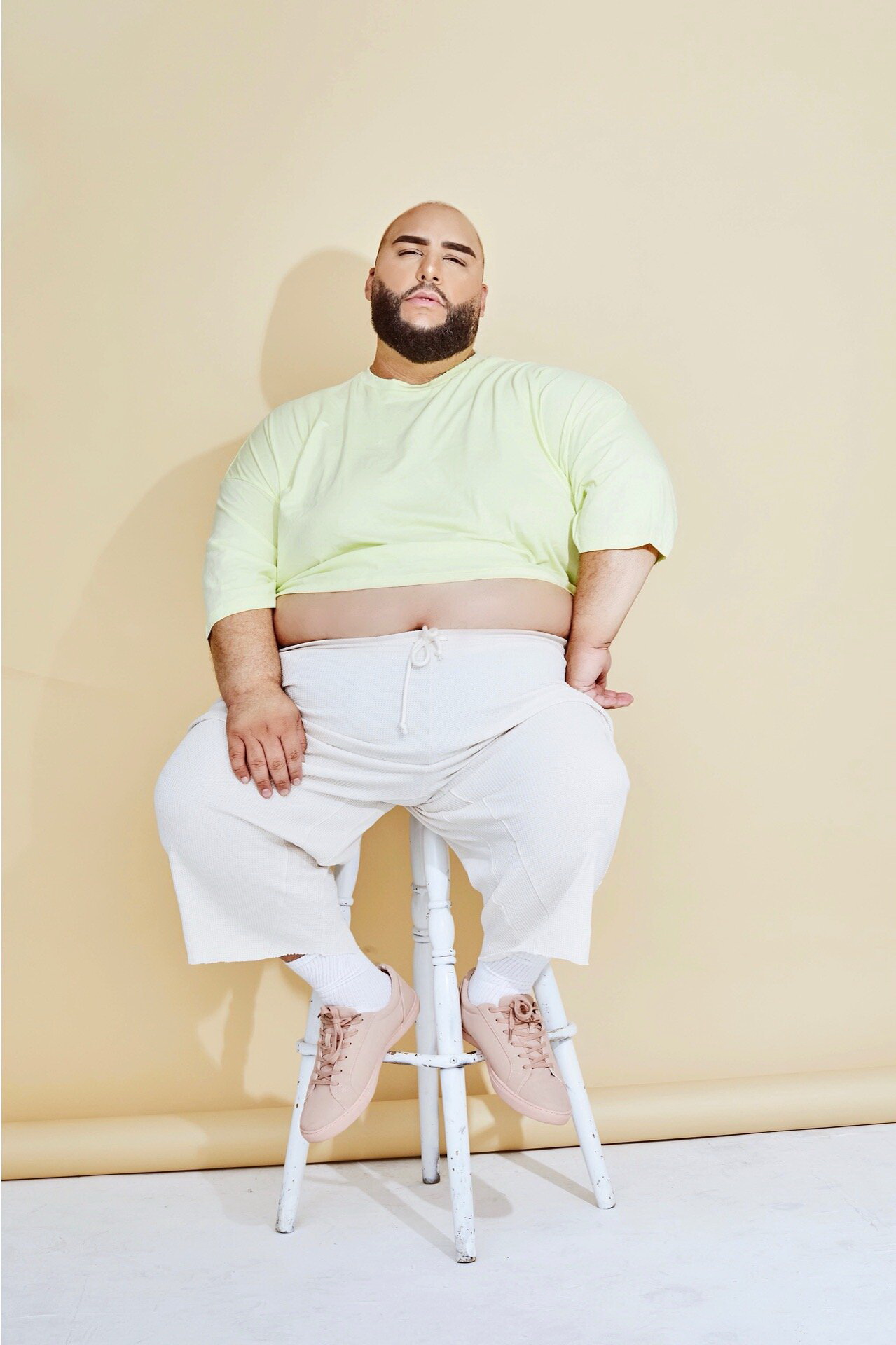 Ady is sitting on a white stool, against a beige wall wearing a mint t-shirt and white loose pants. His belly is partially uncovered. His left arm is bent on his hip and his right arm is relaxed along his body. He is sitting with his legs open. He is wearing white socks and light pink trainers.