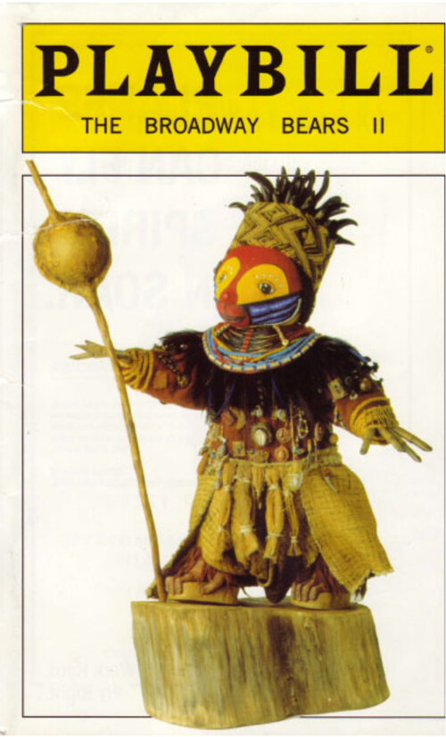 Toy Bear dressed up as Rafiqi from the Lion King. It is inserted in a promo poster with yellow sign saying “Playbill, The Broadway Bears II”. The bear has a red face, is wearing a fabric crown, a colorful collar with black feathers and hanging little mirrors, a fabric skirt and is standing on a wood chop. He is holding a stick with a ball on top.