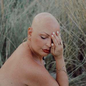 Silvia is a white bald lady. The picture shows her face, part of here back and right shoulder. Her righr hadn covering her left eye. She is wearing some thin golden bracelets, gold ring on her middle finger, and thin round earring on her right ear. Her lips are red and she is looking down. Tall grass in the background.