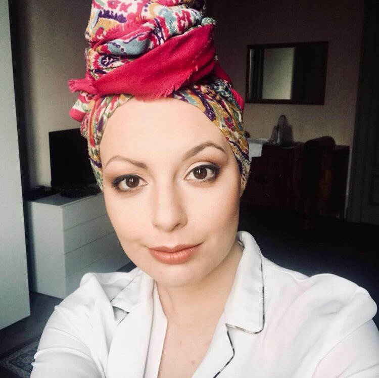 Silvia is staring at the camera wearing a colorful turban. Her brown eyes are wide open, and she is slightly smiling. She is wearing a white shirt with a blazer collar with a darker hem. Details of a living in the back.