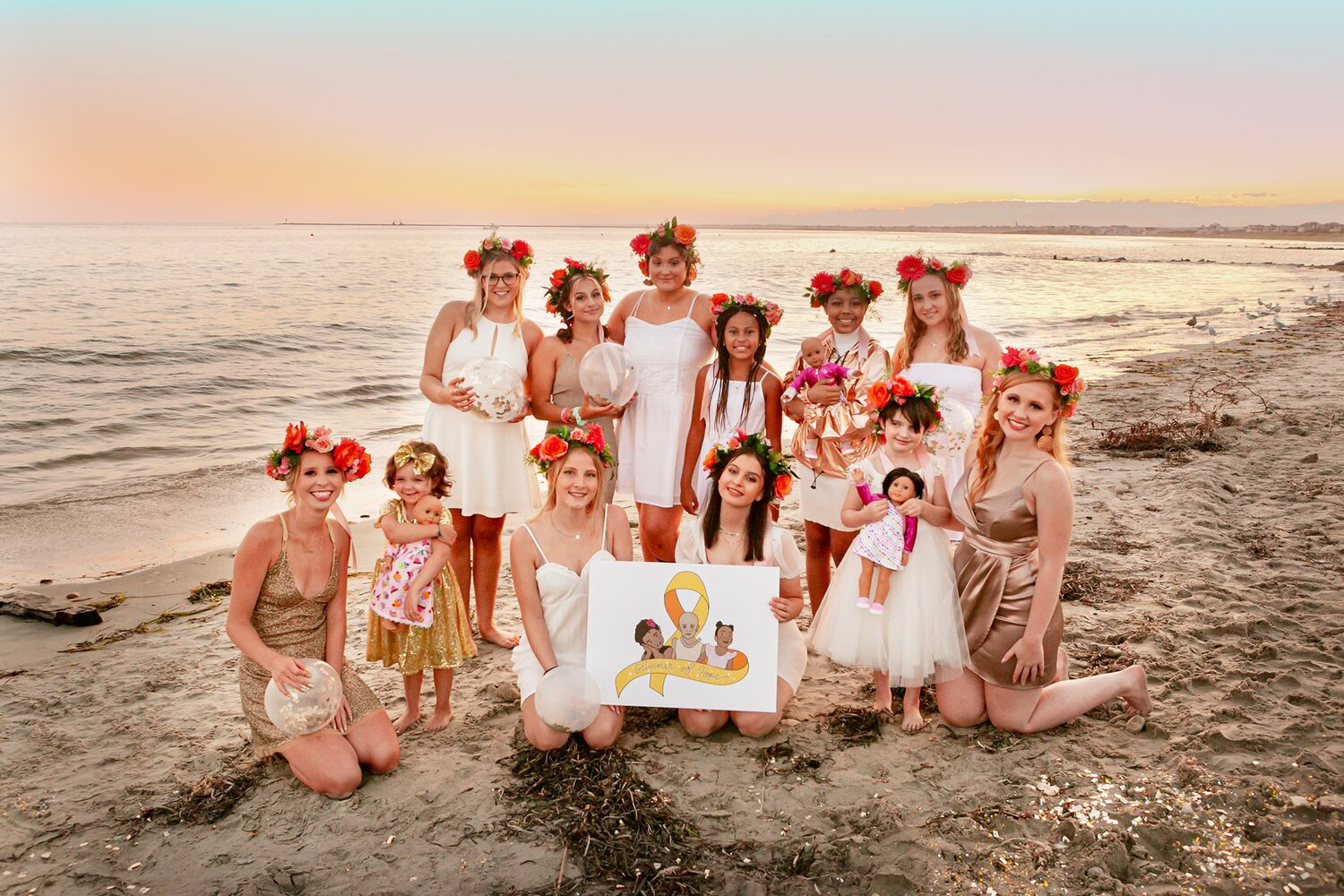 Image of Glimmer of Hope Foundation members at the beach during sunset. They are all girls and are wearing white and gold short dresses, as well as flower arrangements on their hair.