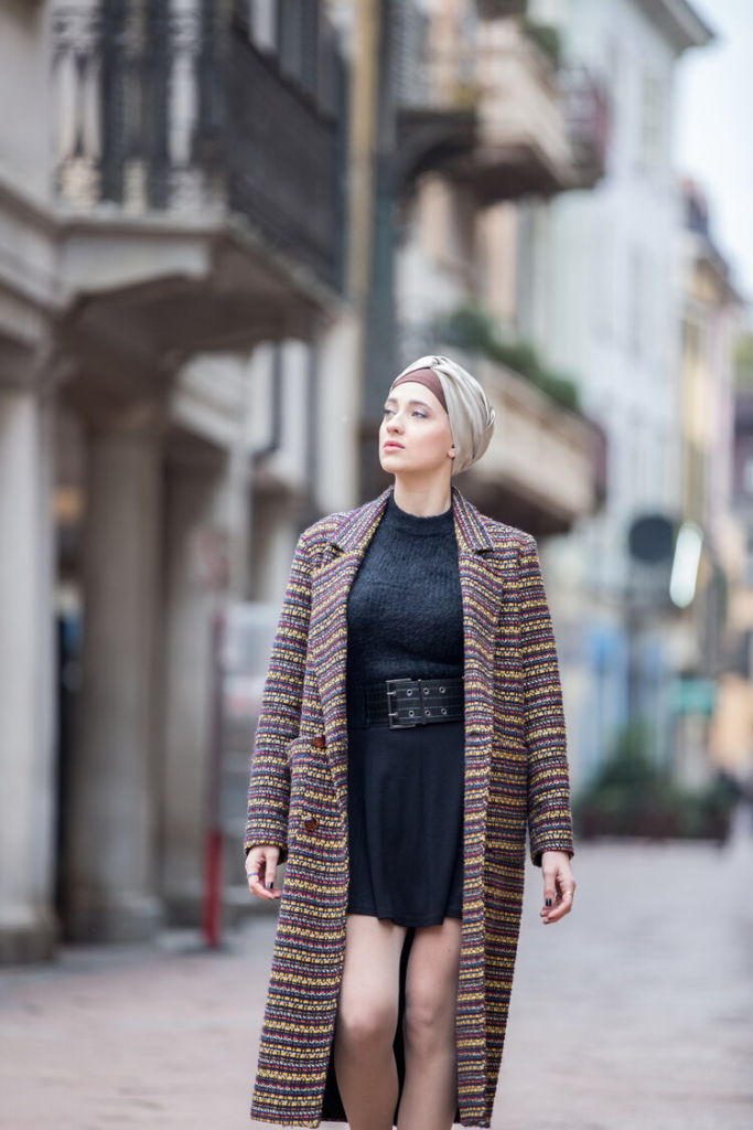 The model is a white young lady walking down the street. She is wearing a short little black dress with a high leather belt. She is wearing a coat, open in the front with horizontal lines in blue, red, yellow. She is obviously wearing a turban with a dark brown cover and lighter brown knotted top.