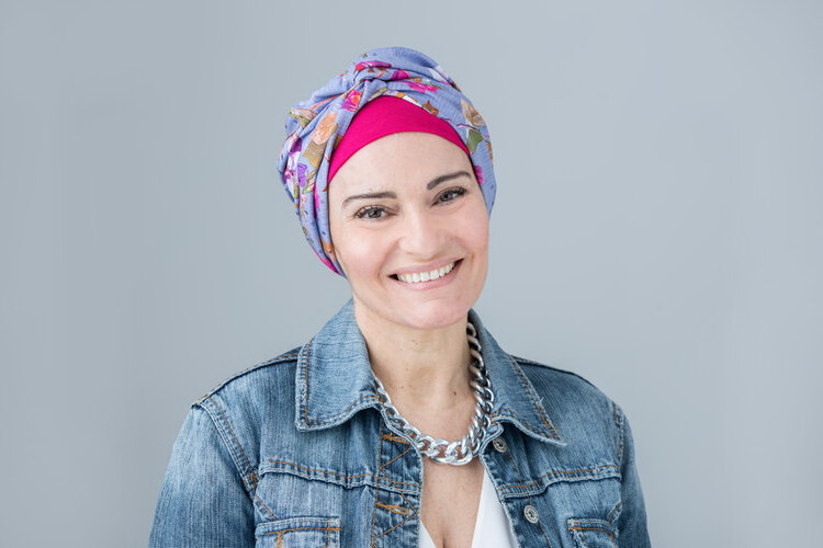 This is a portrait of a white model with dark eyes, wearing a turban in fucsia and completed by a knotted cover with lilac and colorful flowers. She is wearing a denim jacket, white v-neck t-shirt and a chain necklace. The model is smiling to the camera.