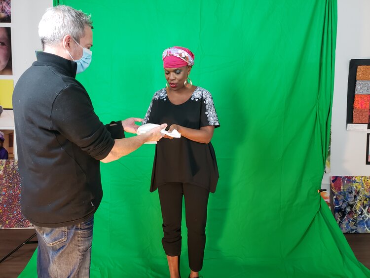 This is a backstage picture from the shooting of the Bigger Plans Project. Rick, standing on the left is sharing something with Zazel, black womand in the centre of the picture. Green screen and some paintings on the back. Rick is wearing a mask, a black sweater and jeans Zazel is wearing a colorful Turban, black t-shirt with white hand-painted shoulders, black pants.
