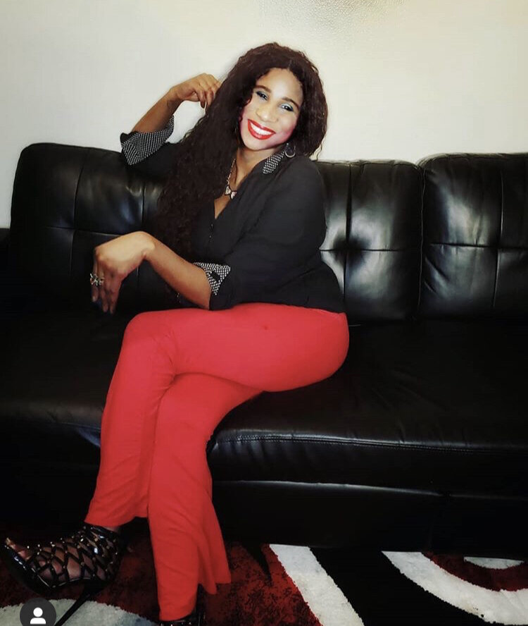 Lachi sitting on a leather black couch, with her legs crossed. She is wearing the black Alana Soul Blouse with White Dots, red pants and black sandals. She is smiling looking at the camera.