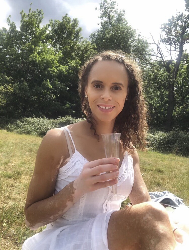 Natalie is sitting on the grass, holding a glass in her right hand. She is wearing a beautiful white dress.