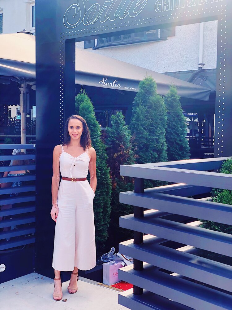 Natalie is standing at the entrance of a cafè, she is wearing a white a summer jumpsuit, nude high heels sandals and she is smiling.