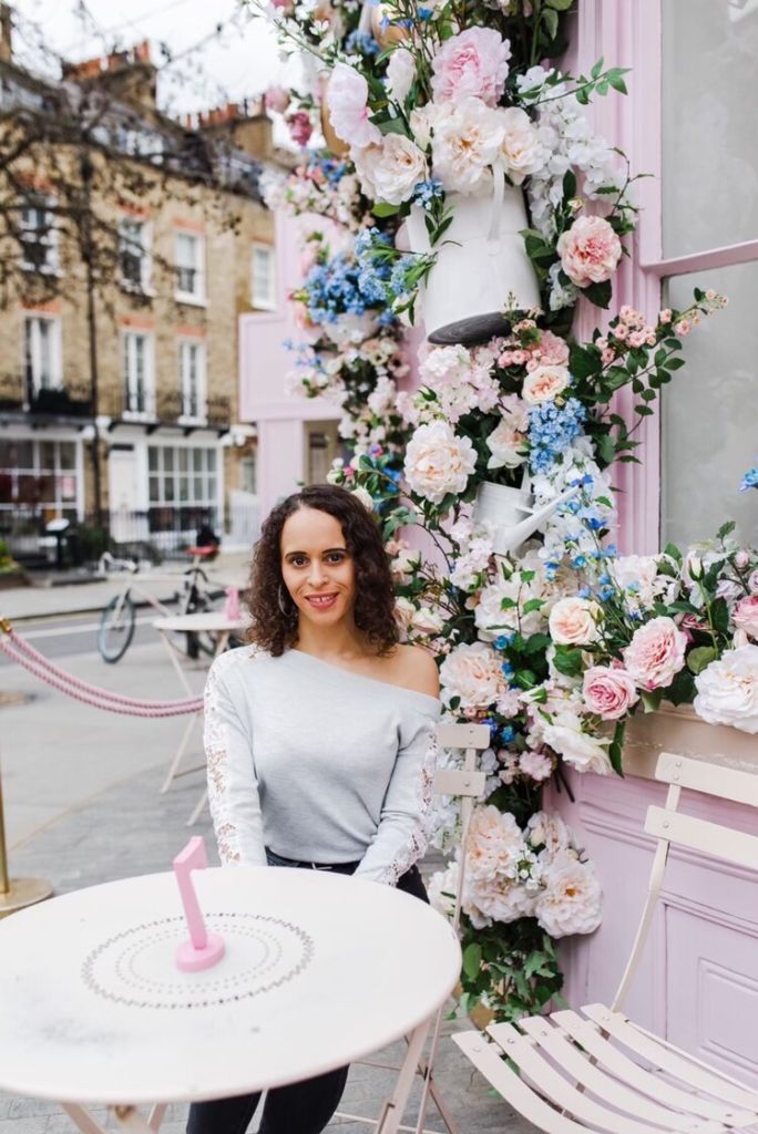 Natalie is sitting at a white round coffee table, with colorful flowers on the wall behind her. She is wearing a grey sweater with laces on her arms and a shoulder out. She is smiling.