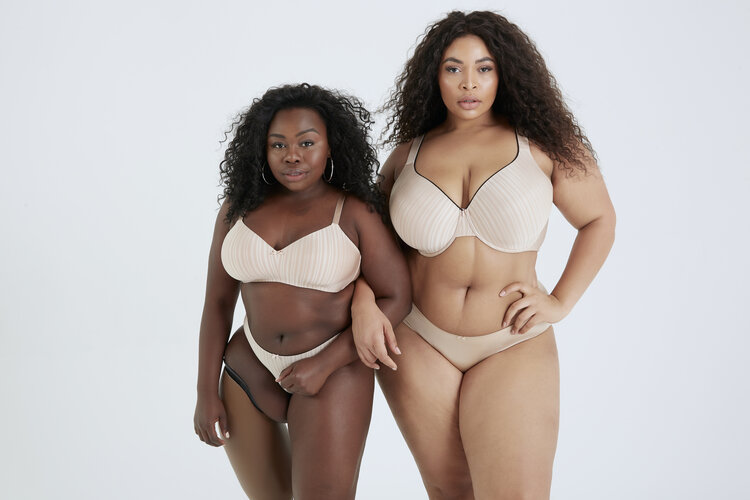 The image shows two women, side by side. The left woman is black, shorter, and wears a prosthesis on her right leg. The woman on the right has a lighter complexion and is taller. The two women have soft curves and wear lingerie that fit each of their bodies perfectly.