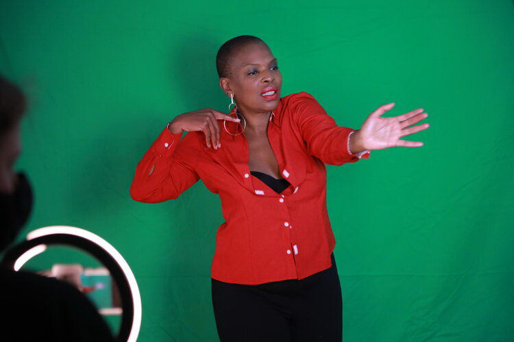 The picture shows Zazel , a black woman with very short hair, wearing a red fitted blouse, The blouse is half open and you can see a black bustier underneath. She is in a dancing pose with her right hand touching her right shulder and her left arm out. Green screen behind her
