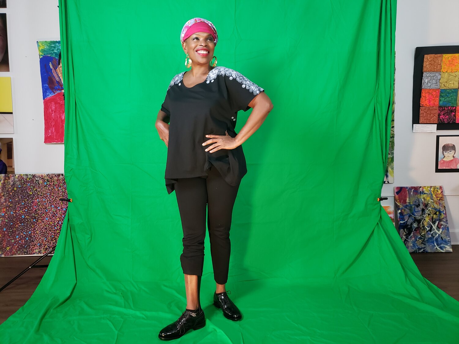 Zazel is standing smiling in front of the green screen at the Positive Exposure location in NYC. She is wearing a black t-shirt with white hand painted shoulders, black pants, black shiny shoes and of course, her fucsia and flowery milk fiber turban