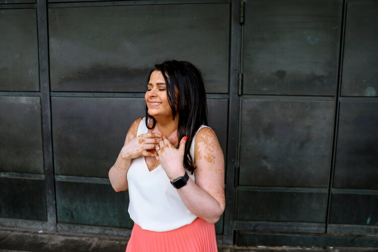 In this photo Joti, a woman with Vitiligo, is standing three quarters turned to her right. The background is a black squared wall. She is in a happy smiling expression, keeping her eyes closed. She has her hands extended by lightly touching the base of her neck. She wears a white sleeveless V-neck top and a pleated coral skirt.