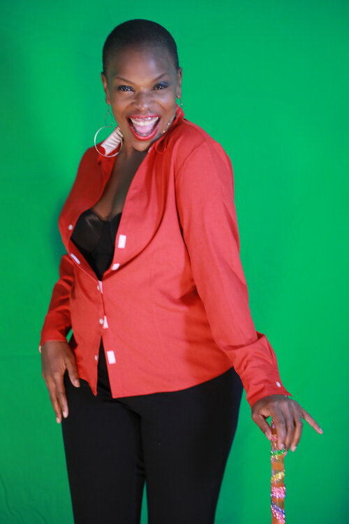 African American woman standing in front of the green screen. She is standing confidently with her face in a radiant expression. She is wearing a fitted red blouse, a balck bustier and black pants. She has her left hand on a colorful walking stick,