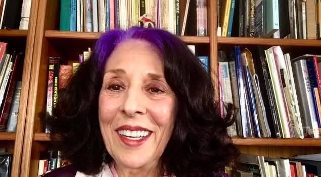 Barbara Beccio, smiling white lady with shoulder length dark brown hair and purple hair roots. Her hair is slightly curly. She is wearing some dark red lipstick and is sitting in front of her book shelves.