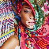 A closeup headshot of model with vitiligo, Winnie Harlow. Her brightly colored silk scarf headdress sweeps up to conceal half of her face.