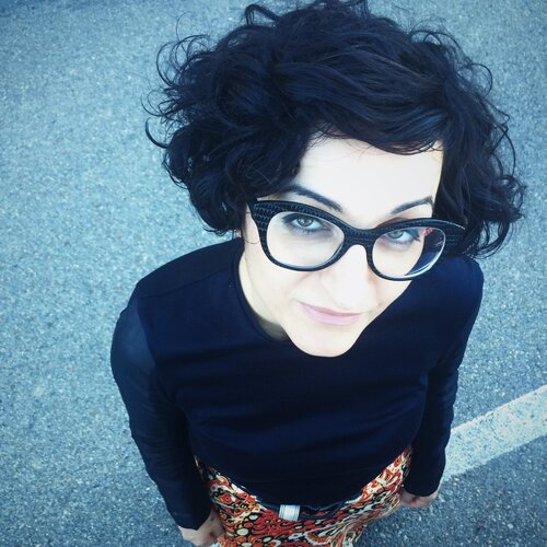 Portrait from above showing Francesca, a white girl with short curly black hair. Francesca wears a pair of black-rimmed butterfly glasses. She is photographed standing on the street, wearing a black turtleneck sweater and a skirt with a patterned print. She has her arms straight down.