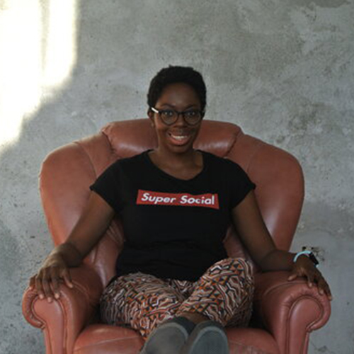 Picture of Sambu Buffa, contributor, black lady smiling with short hair, wearing a black t-shirt with read writing “Super Social” and colorful pants. She is sitting on a sofa.